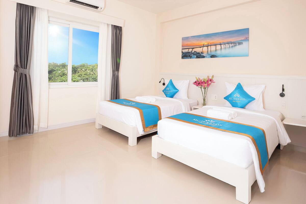 A hotel room with two beds, a large window with curtains, a painting of a pier, and a view of greenery.