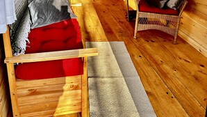 L’intérieur d’un chalet accueillant, avec un plancher en bois, un lit recouvert d’une couverture rouge, une chaise en osier et une petite table avec une lampe.