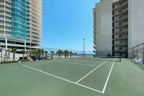 A tennis court with a net, surrounded by a fence, and adjacent to a beach with palm trees.