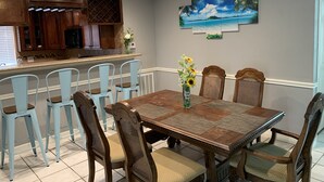 Een eethoek met een houten tafel en stoelen, een keuken op de achtergrond en een schilderij met een strandthema aan de muur.