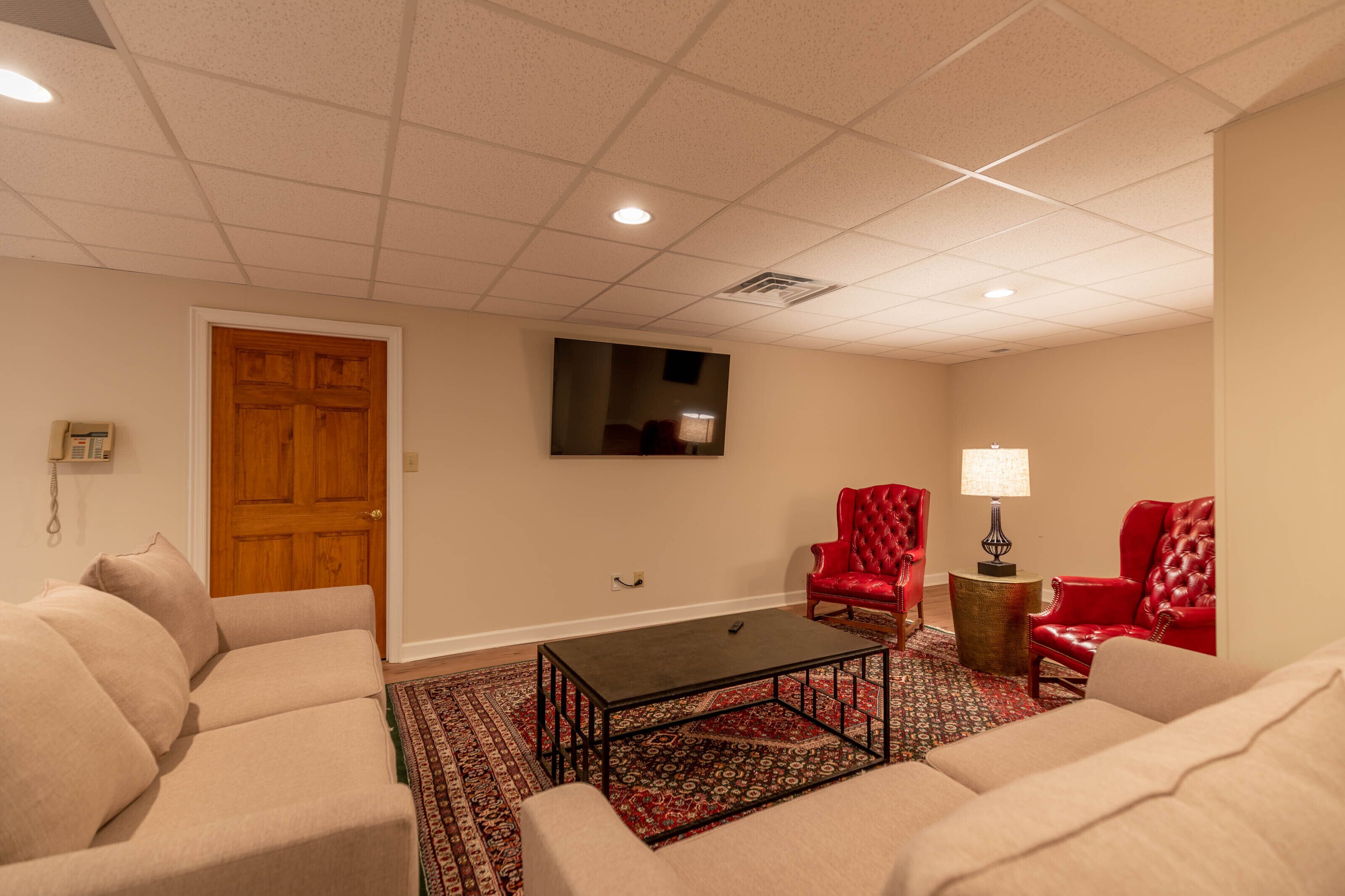 A living room with beige sofas, a coffee table, red armchairs, a TV mounted on the wall, and a patterned rug.