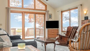Uma sala de estar aconchegante com um sofá, uma mesa de centro e cadeiras de madeira. Uma TV de tela plana está fixada na parede, e há uma grande janela com vista para o exterior.