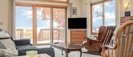 Una sala de estar acogedora con un sofá, una mesa de centro y sillas de madera. Un televisor de pantalla plana está montado en la pared, y hay un amplio ventanal con vistas al exterior.