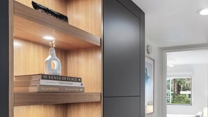 A modern interior with wooden shelves displaying books and a crystal. A grey cabinet is visible, and a bedroom with a bed and window is in the background. - Beautifully designed with Mountain View, Resort Amenities, Golf, Office + More (Palm Springs)