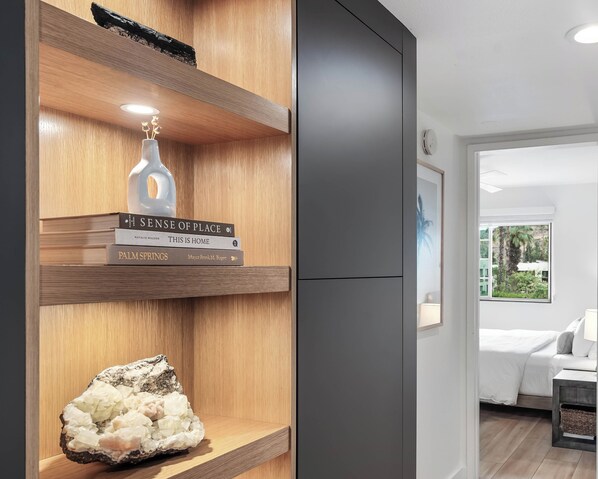 Ein modernes Interieur mit Holzböden, auf denen Bücher und ein Kristall ausgestellt sind. Ein grauer Schrank ist sichtbar, und im Hintergrund befindet sich ein Schlafzimmer mit Bett und Fenster.