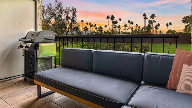 A balcony with a black leather sofa, a grill, and a view of palm trees and a sunset. - Beautifully designed with Mountain View, Resort Amenities, Golf, Office + More (Palm Springs)