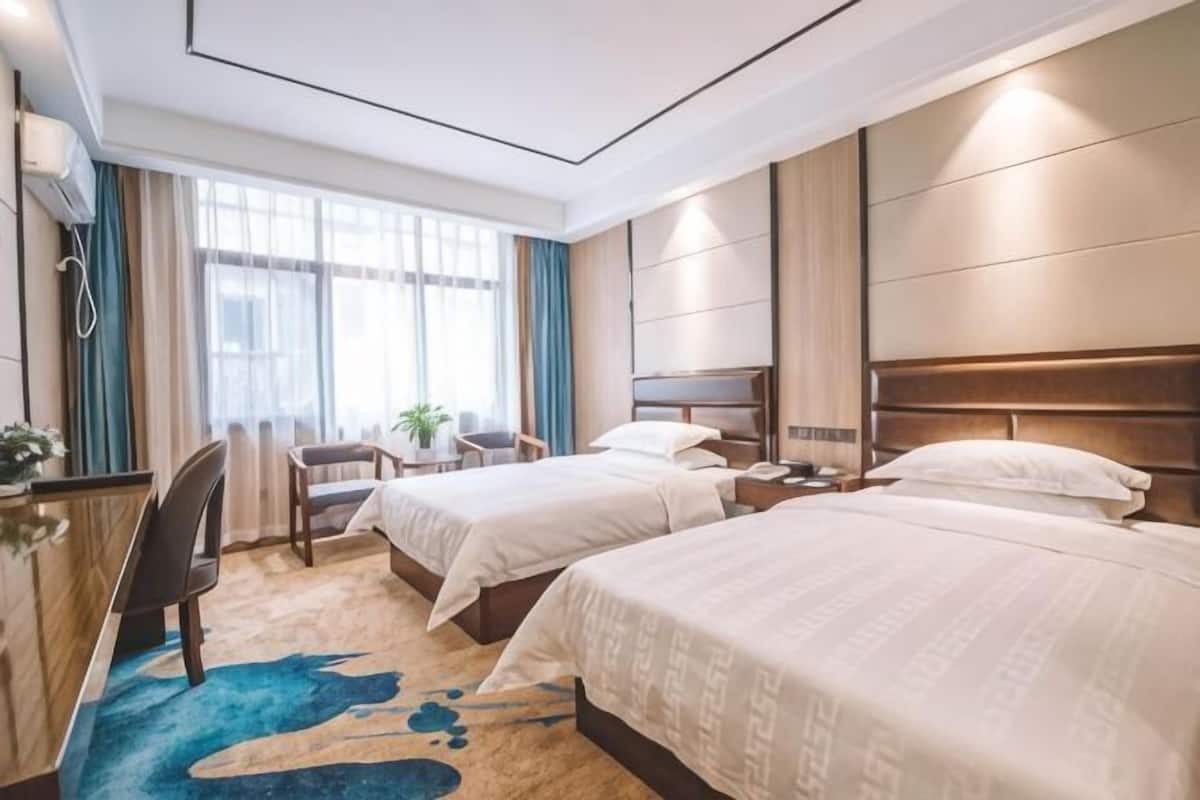 Quarto de hotel com duas camas, uma escrivaninha, uma cadeira, uma mesa pequena e uma janela com cortinas.