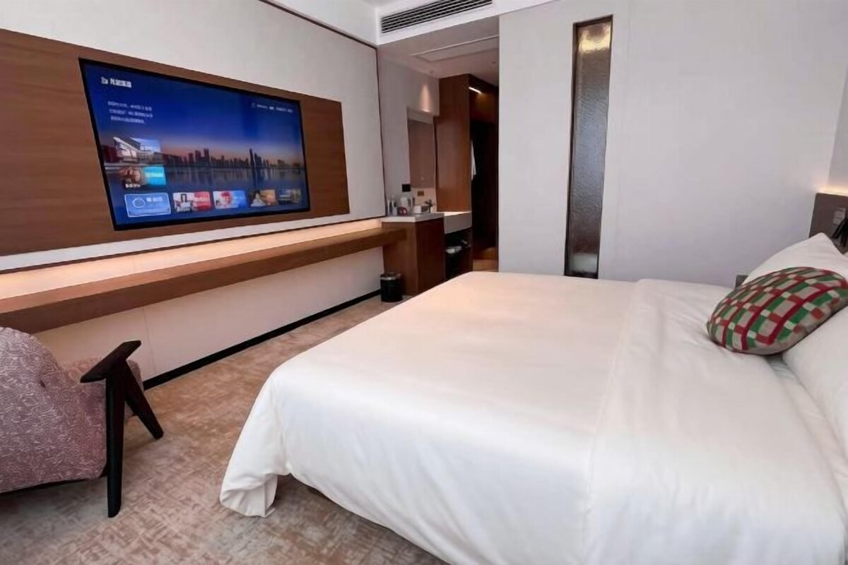 Ett hotellrum med en stor platt-TV som är monterad på väggen, en säng med vita sängkläder och en liten sittgrupp med en stol och en kudde.