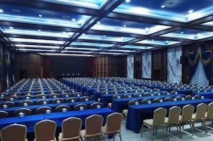 Un salón de conferencias con filas de mesas y sillas, un techo alto con iluminación y un escenario al frente.