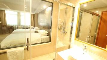 A hotel room with a bed, a glass-enclosed shower, a sink, and a large mirror.