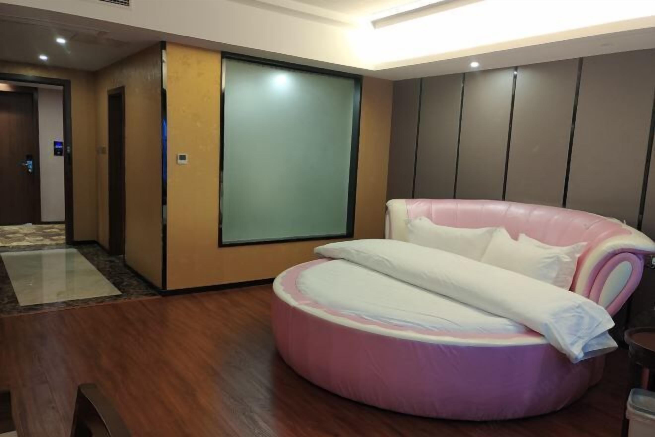 A modern hotel room with a wooden floor, a pink and white circular bed, a large sliding door, and a view of the hallway.
