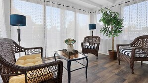 Ein Zimmer mit Korbmöbeln, einem Glastisch und einem großen Fenster mit durchsichtigen Vorhängen.