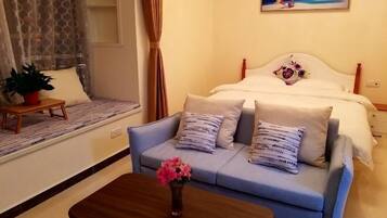 A bedroom with a bed, a sofa, a small table with a vase of flowers, and a window with curtains.
