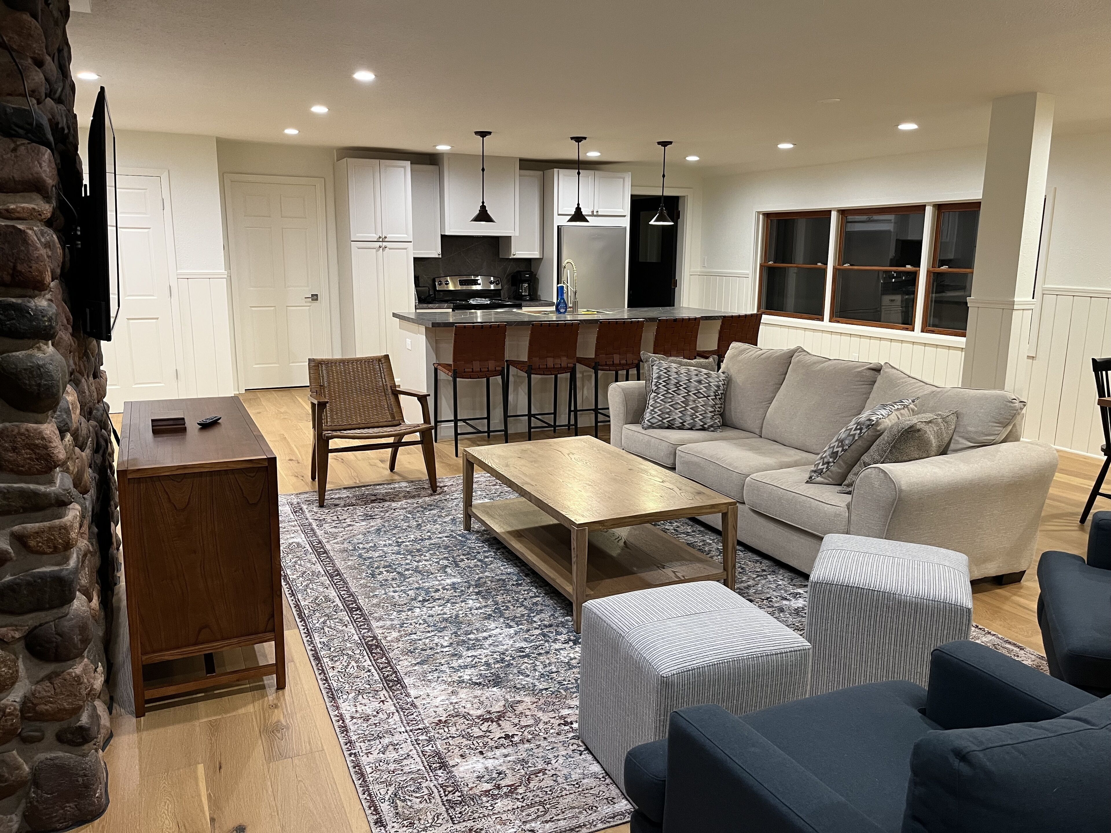 A modern living room with a sofa, ottoman, coffee table, and a rug. There is a kitchen area with a refrigerator, stove, and bar stools.