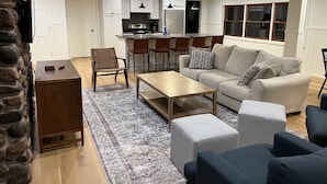 A modern living room with a sofa, ottoman, coffee table, and a rug. There is a kitchen area with a refrigerator, stove, and bar stools. - 4 Season Fun on Lake Namakagon - Snowmobiling, Boating, Fishing, UTV & More! (Cable)