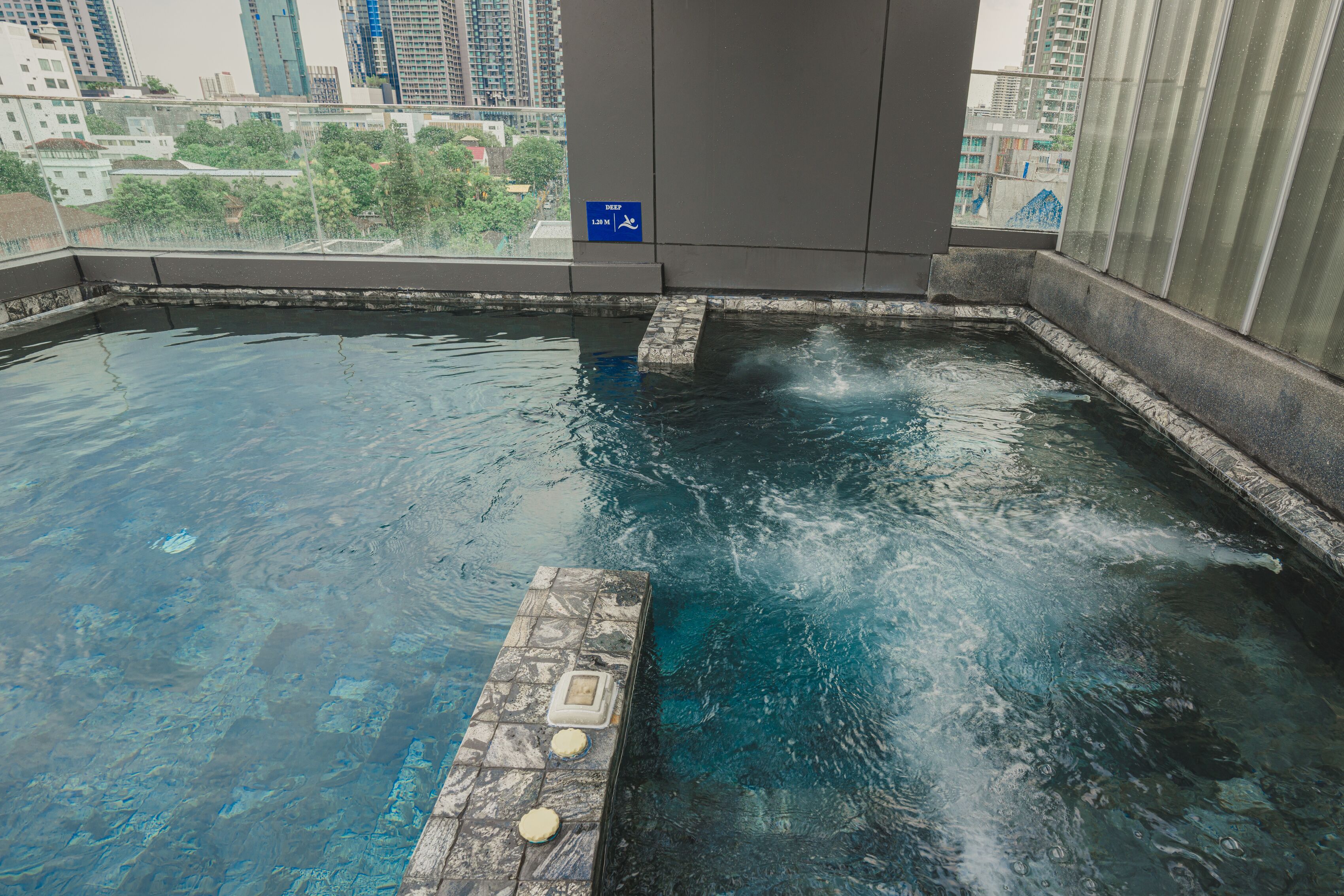 A rooftop pool with clear water, a stone deck, and a view of city buildings.