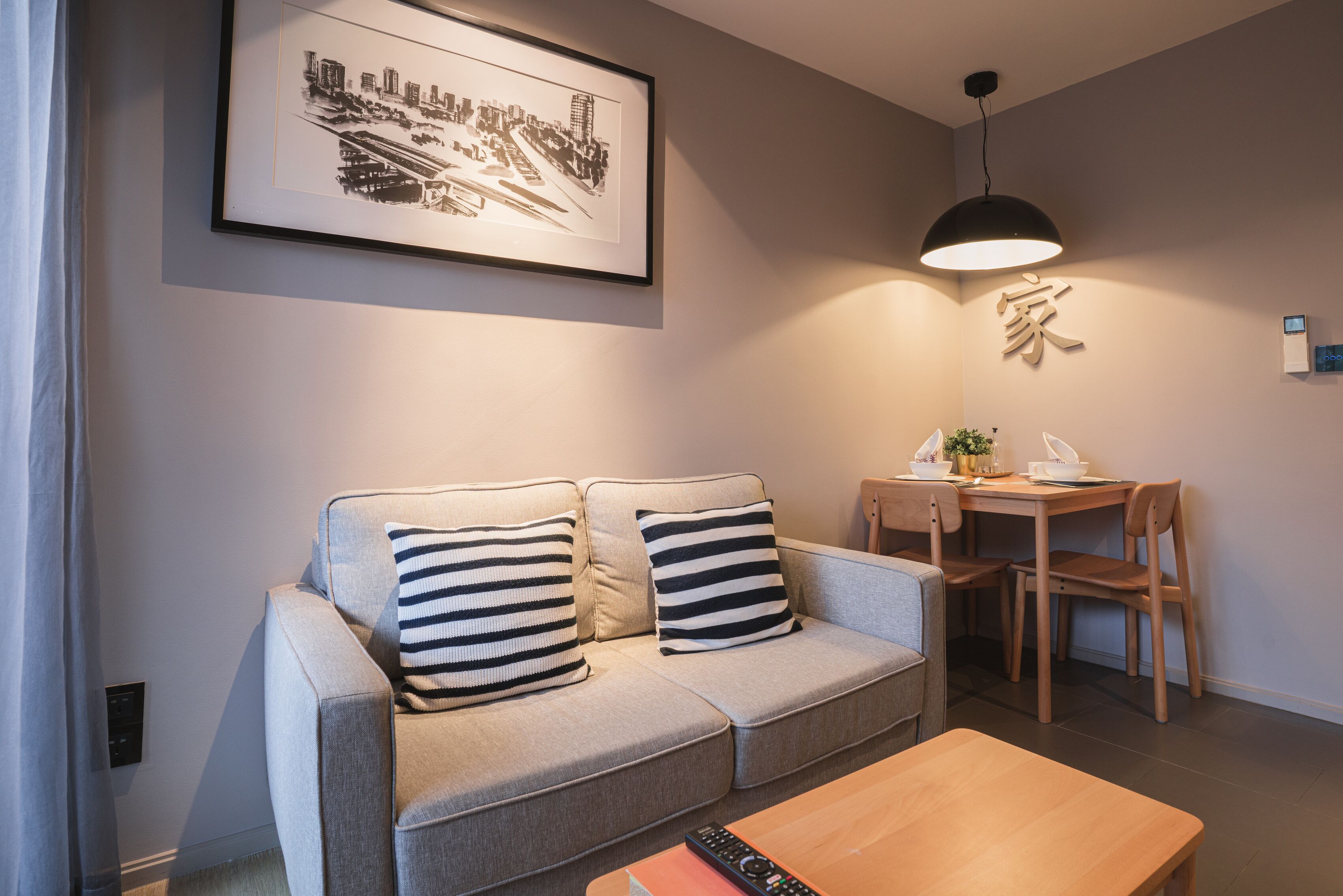 Ein modernes Wohnzimmer mit einer Couch, gestreiften Kissen, einem Couchtisch, einem Esstisch mit Stühlen, einer Pendelleuchte und einem Stadtbild im Bilderrahmen an der Wand.