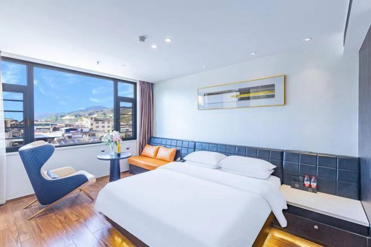 Habitación de hotel con una cama grande, un rincón de descanso con una silla y una mesa, vistas de la ciudad a través de ventanas amplias y una obra de arte enmarcada en la pared.