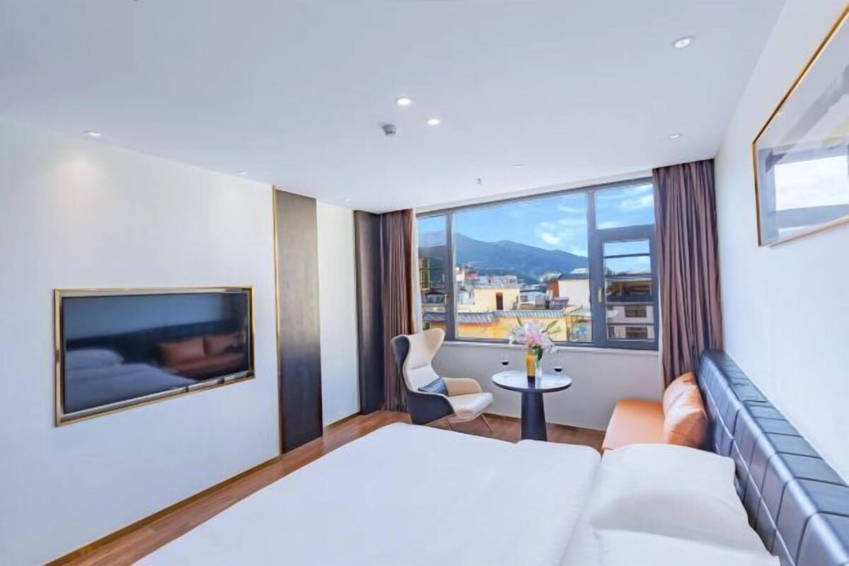 Habitación de hotel con cama, televisor, zona de estar con mesa y un amplio ventanal con vistas a edificios y montañas.