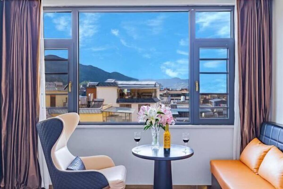 Una habitación con vistas a edificios y montañas, una mesa redonda con un florero y dos copas de vino, un sofá y una silla.