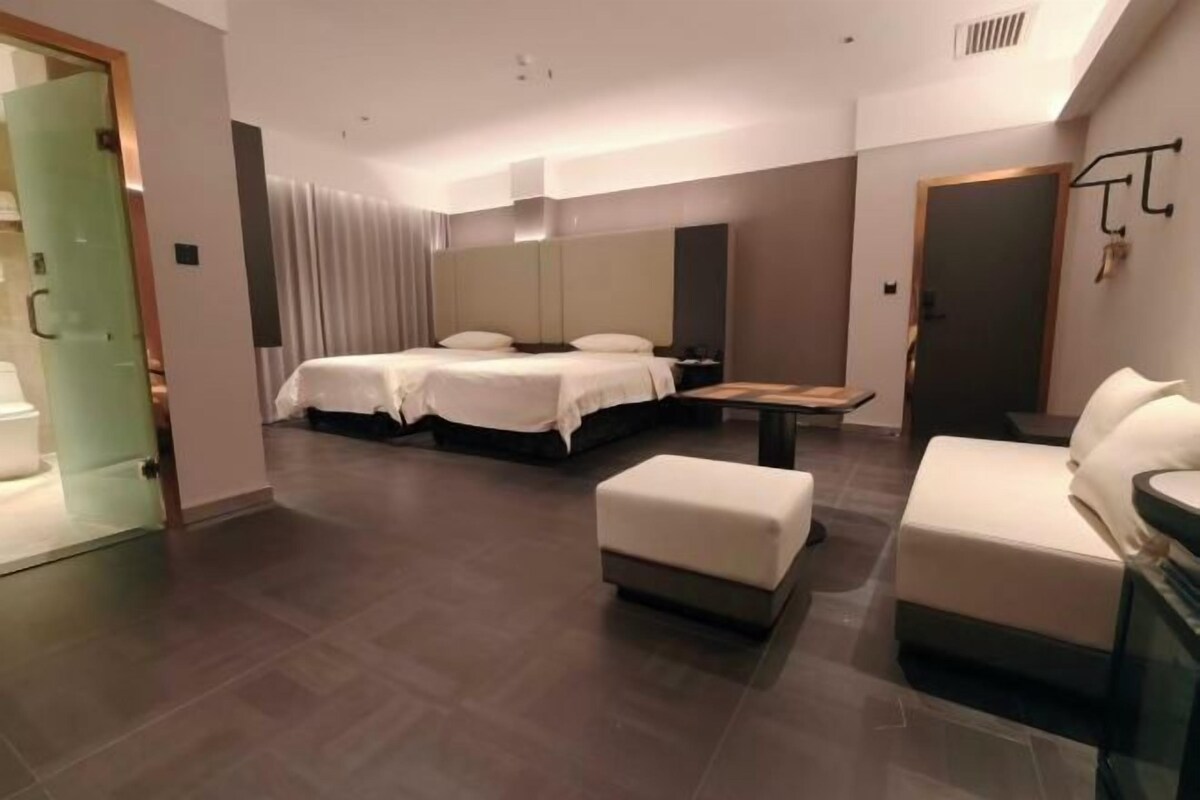 Een moderne hotelkamer met een groot bed, een kleiner bed, een bank, een tafel en een glazen scheiding.