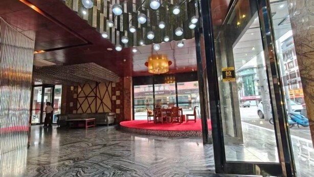 En hotelllobby med ett marmorgolv, ett område med röd matta där det står ett bord och några stolar, och hängande lampor.