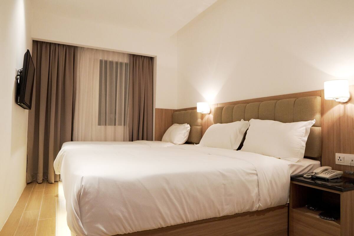 A hotel room with a large bed, a flat-screen TV mounted on the wall, a bedside table with a phone and remote control, and a window with curtains.