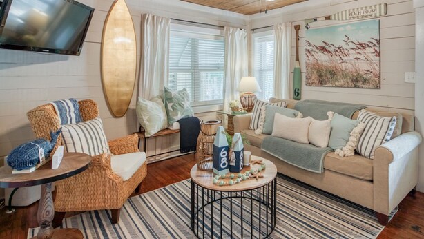A cozy living room with a wooden ceiling, a surfboard, a television, and a beach-themed painting. - Cozy Tybee Getaway on Butler Ave (Tybee Island)