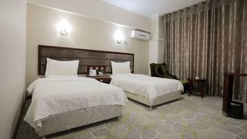 Quarto de hotel com duas camas, uma mesinha de cabeceira, uma cadeira e uma janela grande com cortinas.