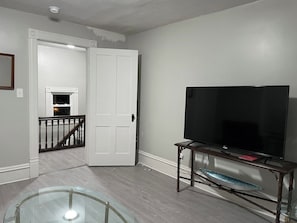 A living room with a television on a wooden stand, a glass table, and a staircase leading to another room. - The Yellow-Shuttered House apt B (Sharon)