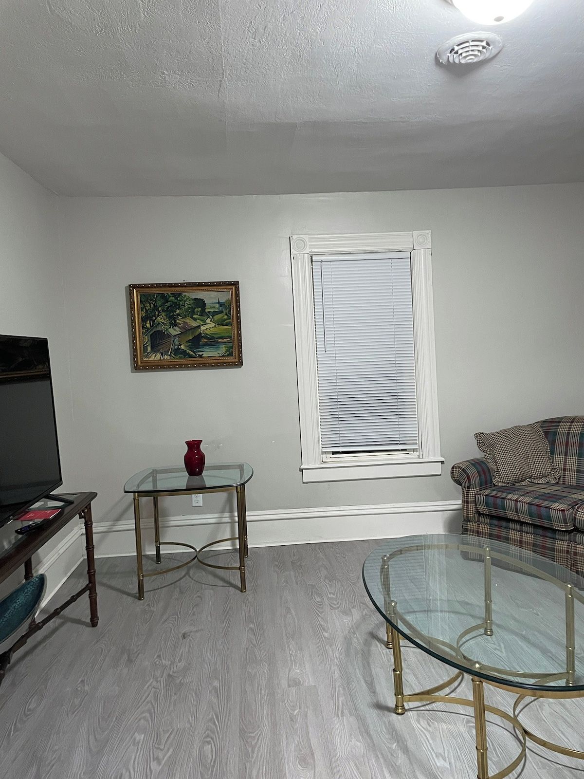 Una sala de estar con televisor, una mesa de centro de vidrio, un sofá, un cuadro y una ventana con persianas.