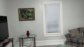 Una sala de estar con televisor, una mesa de centro de vidrio, un sofá, un cuadro y una ventana con persianas.