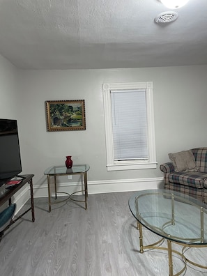 A living room with a television, a glass coffee table, a sofa, a painting, and a window with blinds. - The Yellow-Shuttered House apt B (Sharon)