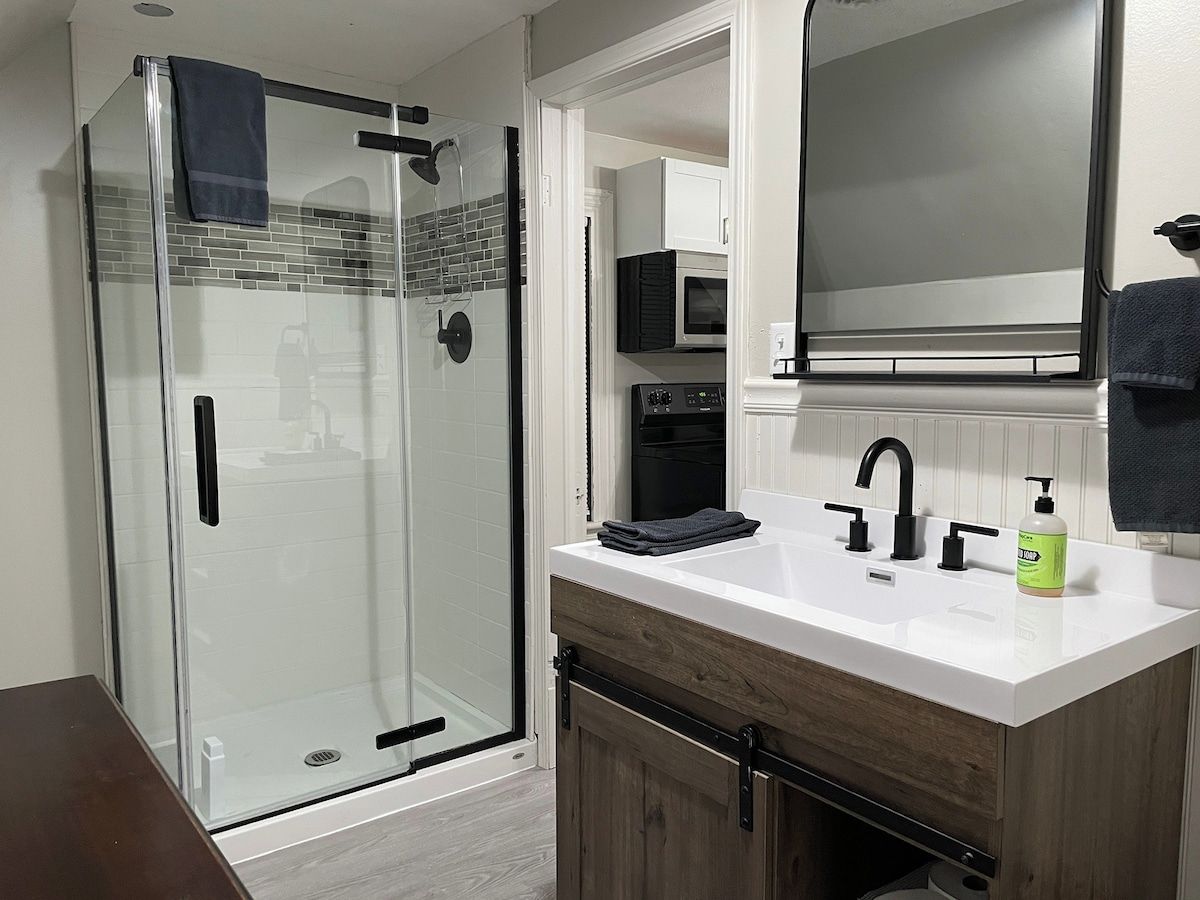 Un baño moderno con cabina de ducha de vidrio, un lavabo blanco con tocador de madera y un espejo grande sobre el lavabo.