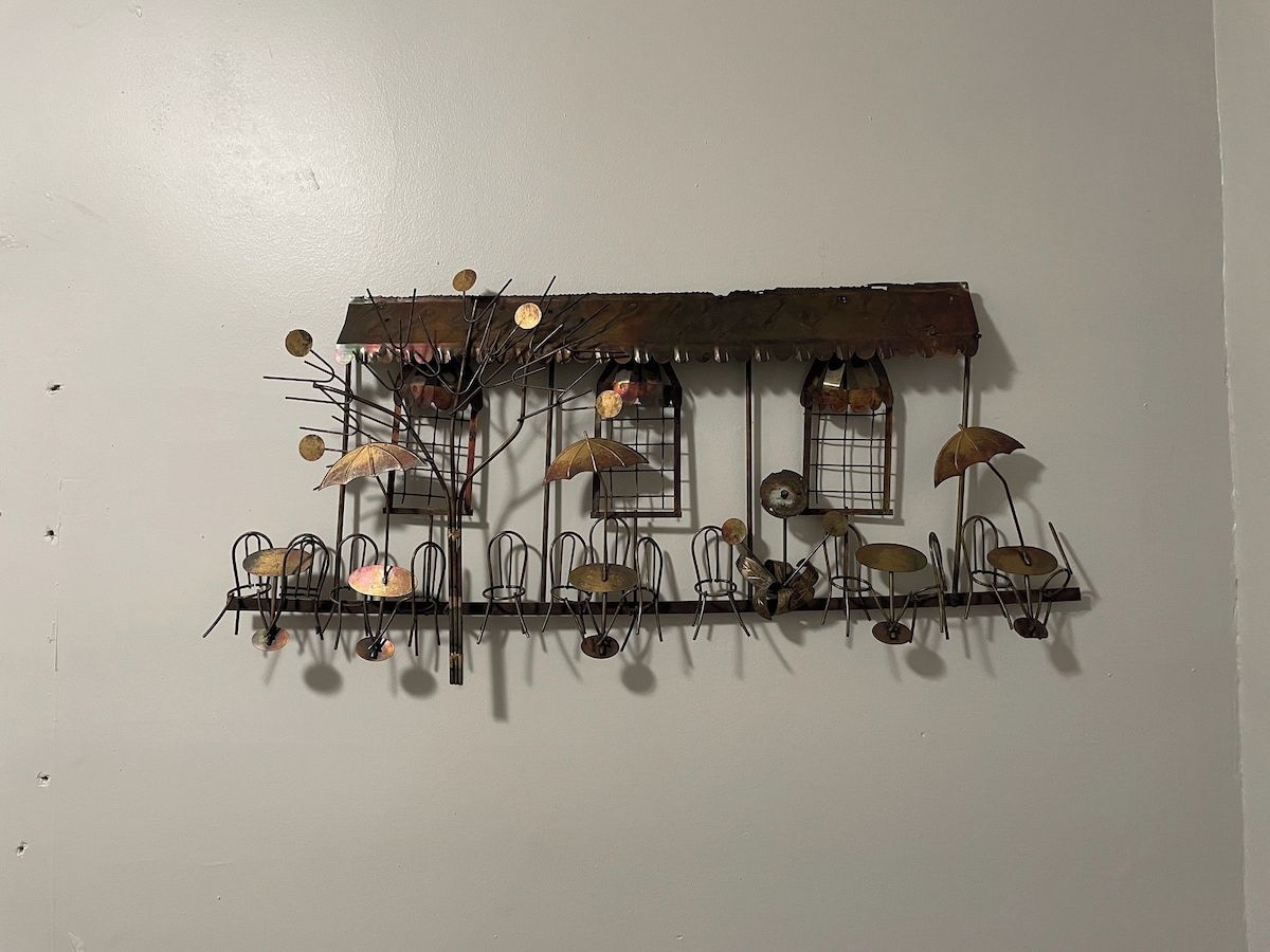 Una instalación artística en metal que muestra una casa rústica con porche, sillas y sombrillas.