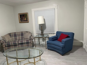 A living room with a plaid sofa, a blue armchair, a glass coffee table, a lamp, and a framed picture on the wall. - The Yellow-Shuttered House apt B (Sharon)