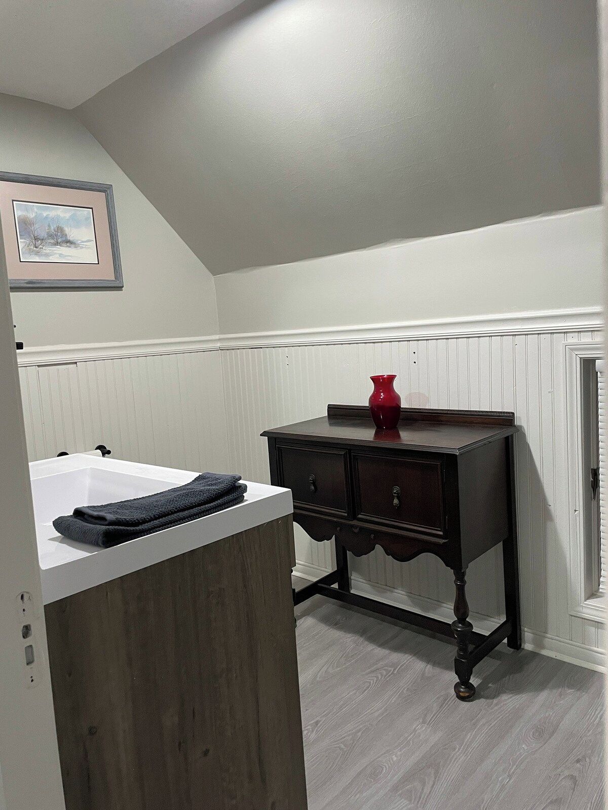 A bathroom with a white bathtub, wooden vanity, and a red vase on the vanity.