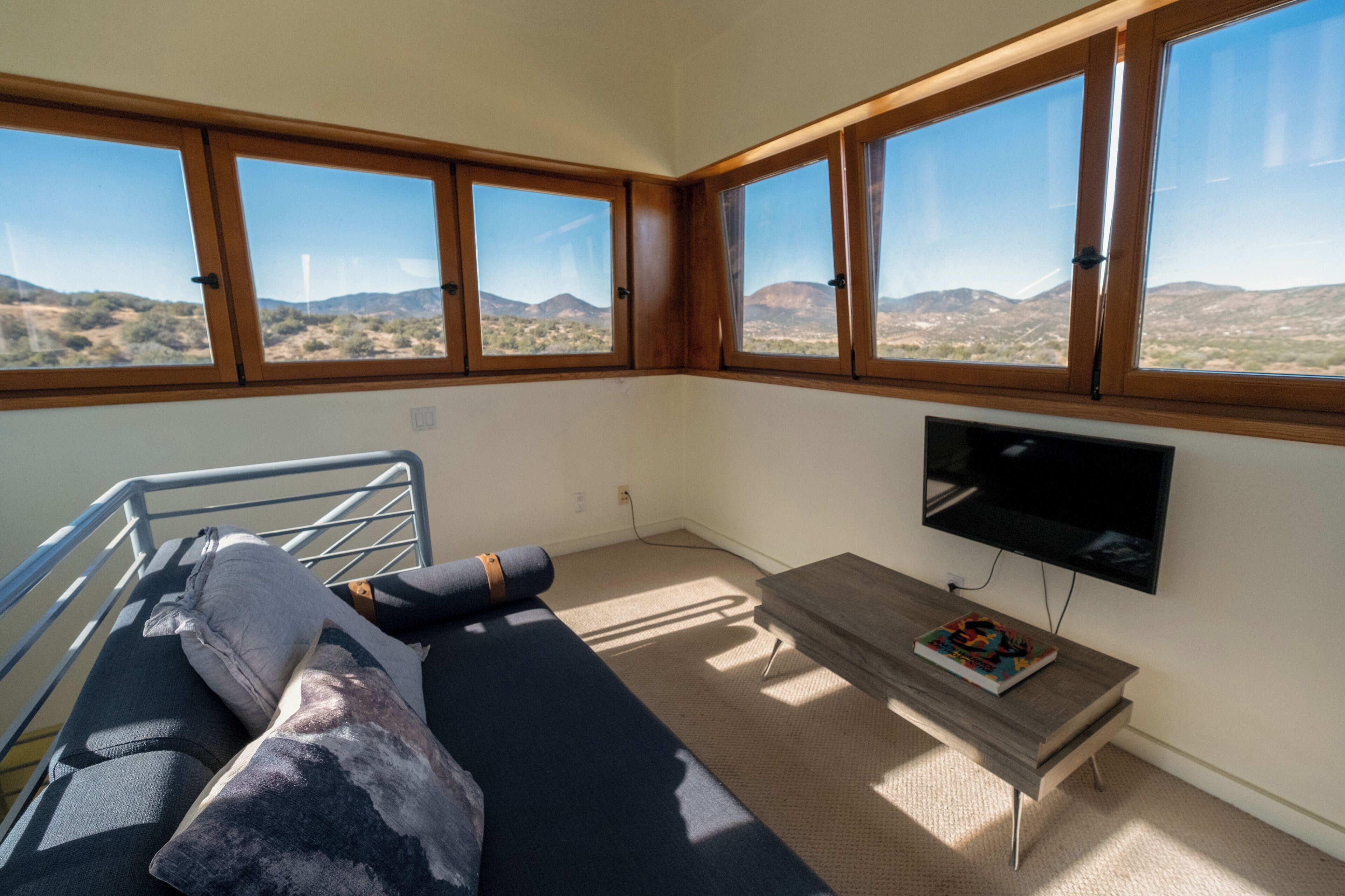 A room with a sofa, a small table, a TV, and large windows with a view of hills.