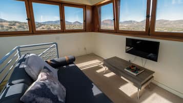 A room with a sofa, a small table, a TV, and large windows with a view of hills.