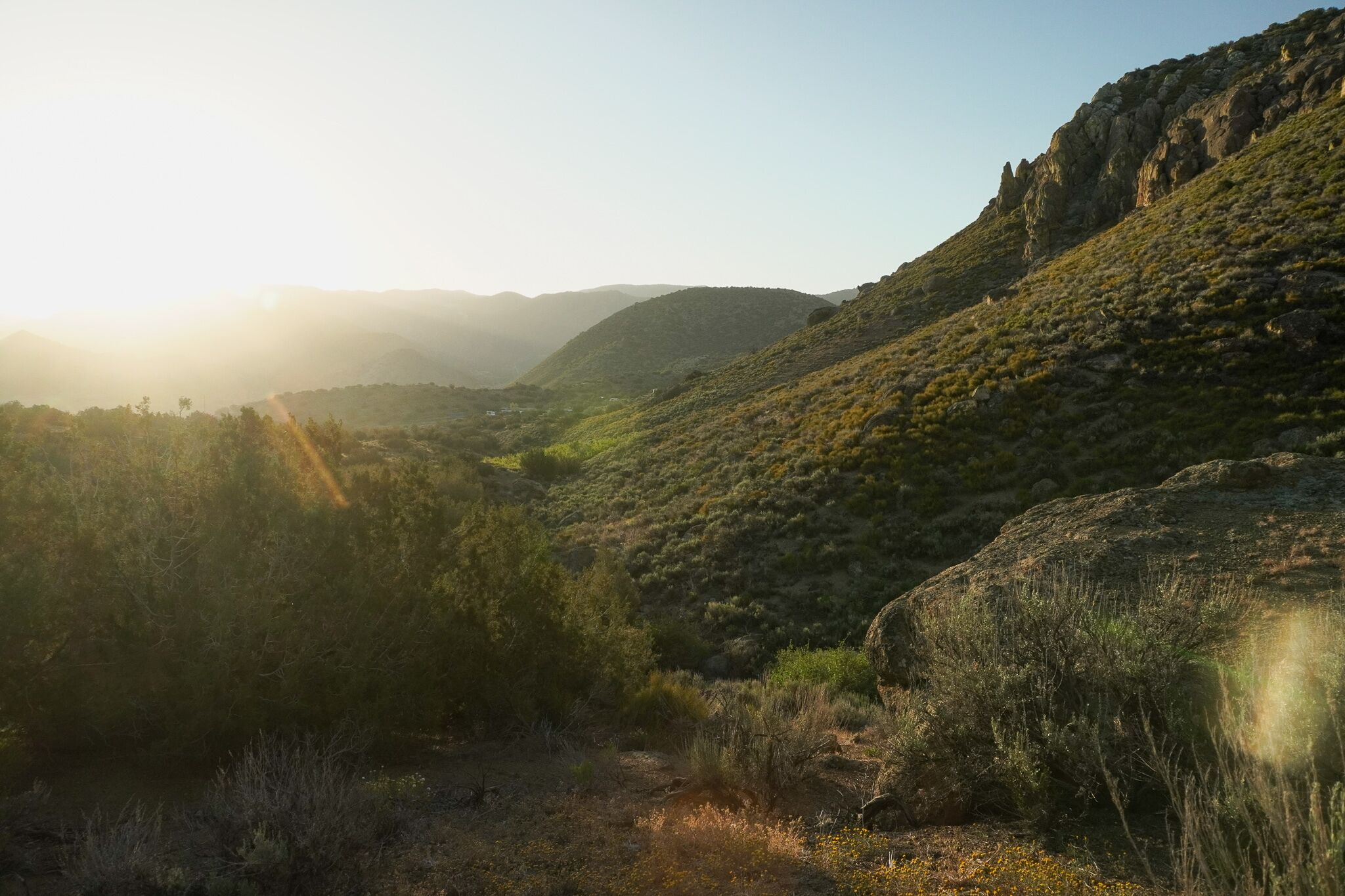Un paysage montagneux, avec un ciel dégagé et la lumière du soleil filtrant à travers les arbres.