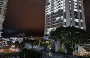 Korkea kerrostalo, jossa on useita kerroksia ja ikkunoita, ja sen ympärillä on puita sekä pieni silta.