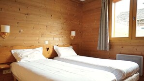 A wooden cabin room with two beds, each with white bedding, a window with curtains, and wall-mounted lamps.
