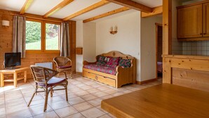 A cozy room with a wooden bench, a television, wicker chairs, a sofa with patterned cushions, and a kitchen area with wooden cabinets.