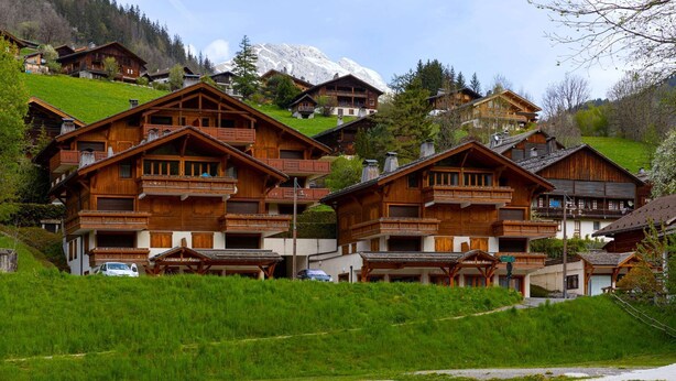 A cluster of wooden chalets with balconies, set against a backdrop of green hills and snow-capped mountains.