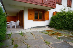 Eine terrasse mit Steinplatten, weißen Stühlen und einem Holztisch, ein weißes Gebäude mit Holzläden und ein grüner Busch.