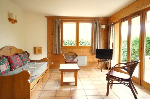 Ein gemütliches Zimmer mit einer Holzdavonia, einem kleinen Tisch, einem Korbstuhl, einem Fernseher und großen, mit Vorhängen versehenen Fenstern.