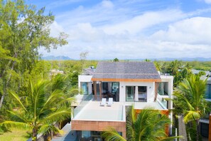A modern house with a glass balcony, surrounded by palm trees and mountains in the background.