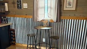 Una stanza accogliente con soffitto in legno, una finestra con tende, un tavolino rotondo con due sedie, un tappeto decorato e una mensola a parete con bicchieri e un piccolo oggetto decorativo.