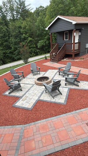 A patio with Adirondack chairs, a fire pit, and a small building with a deck.