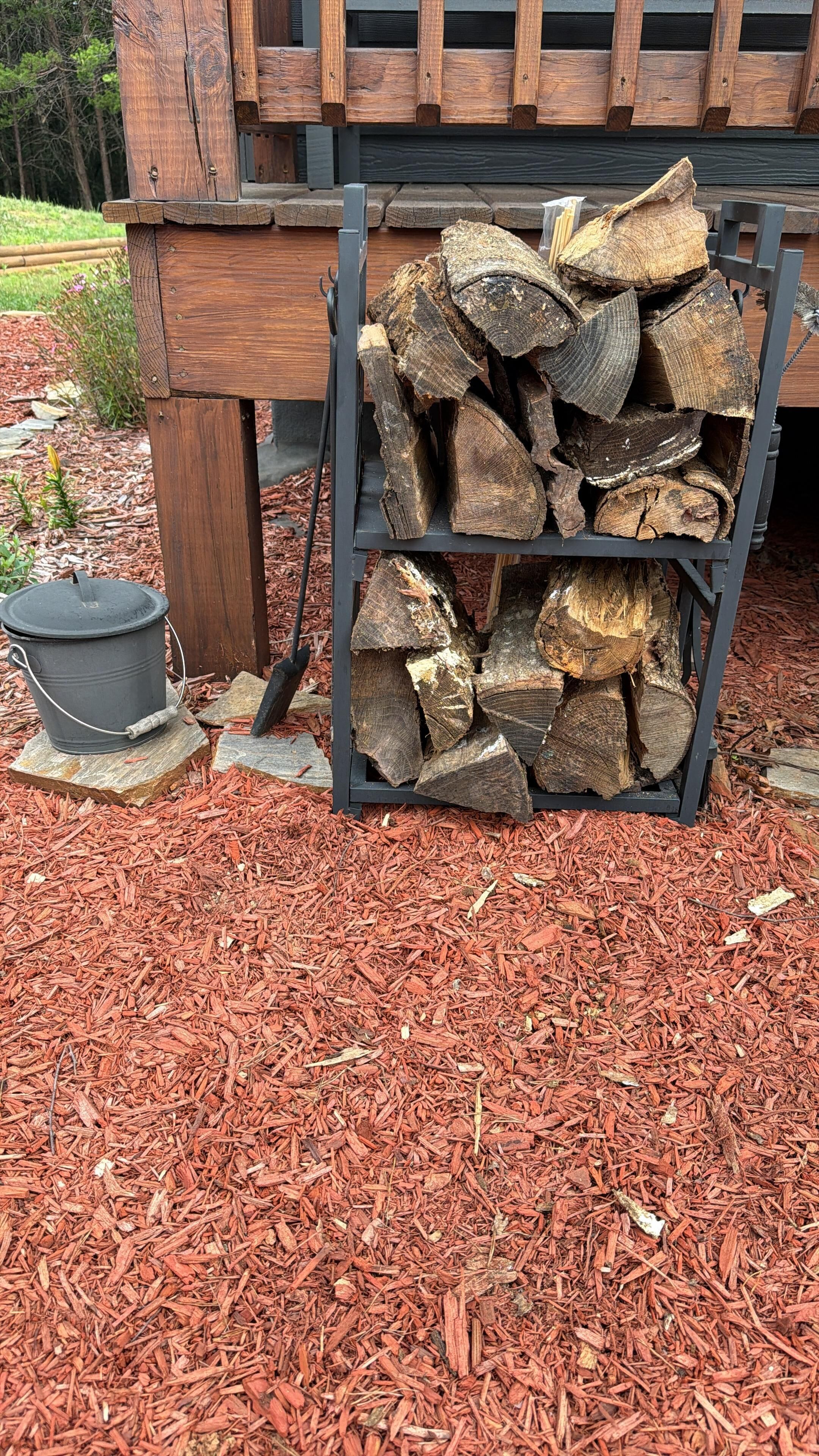 A stack of firewood on a metal rack outside a building with a wooden bench and a metal pot nearby.